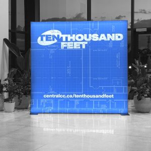 A blue illuminated sign reads TENTHOUSAND FEET, with a blueprint design in the background, displayed indoors near large windows and surrounded by potted plants. A website link is shown at the bottom.