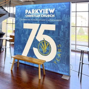 A large blue banner reads Parkview Christian Church and 75 Years with a church logo, celebrating 75 years of God's faithfulness. A wooden bench and two tall round tables sit in front in a bright, modern room.