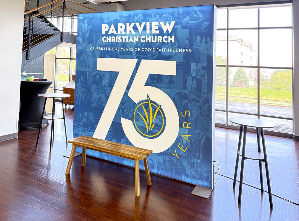 A large blue banner reads Parkview Christian Church and 75 Years with a church logo, celebrating 75 years of God’s faithfulness. A wooden bench and two tall round tables sit in front in a bright, modern room.