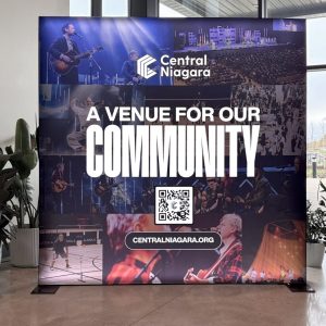 A large display sign reads Central Niagara: A Venue for Our Community with event photos, a QR code, and the website CENTRALNIAGARA.ORG on it. The sign stands indoors next to green plants.