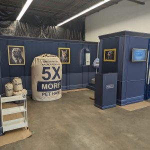 A blue booth in a warehouse features dog portraits, a TV, a hand sanitizer dispenser, and a giant fake lint trap labeled “5X MORE PET HAIR,” along with bags labeled “MAYTAG.”.