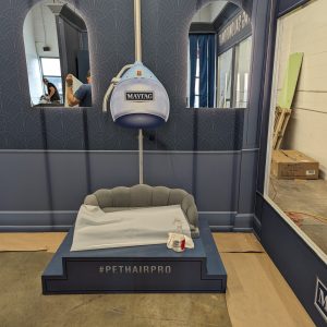 A pet-sized salon setup with a small gray couch, dryer labeled Maytag, and #PETHAIRPRO sign. Mirrors and blue paneled walls surround the area; a person stands partially visible on the left.