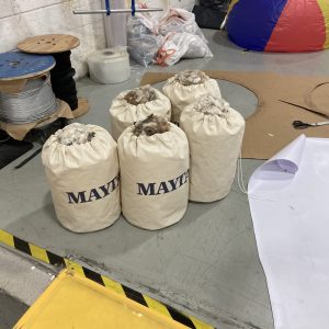 Five canvas bags labeled MAYTI filled with material sit on a gray floor near cables, cardboard, scissors, and a large colorful ball in an industrial setting.