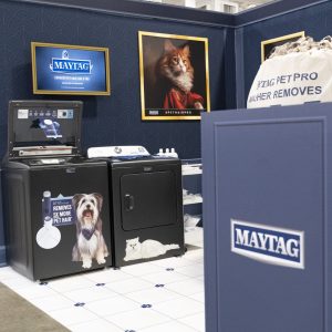 A Maytag appliance display features a washer, dryer, pet portraits, and a large bag of laundry. There are stickers of a dog and cat on the appliances, highlighting pet hair removal. The setup is in a well-lit showroom.