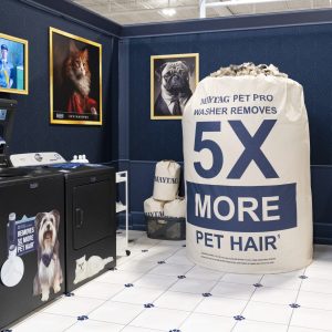 A laundry room display features a washer, dryer, large sack labeled 5X more pet hair, smaller sacks, and framed portraits of cats and dogs dressed in suits hanging on the navy blue walls.