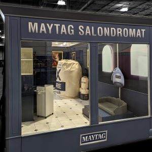 A blue Maytag-themed booth resembling a laundromat with a large laundry bag, cart, and bench, set up inside an event space. The sign above reads MAYTAG SALONDROMAT.