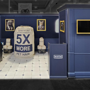 A blue exhibition booth with 5X MORE PET HAIR on a large bag, laundry machines, framed pet portraits, two beige chairs, and blue curtains. TVs are mounted on the walls; the floor is tiled white.