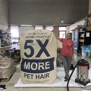 A woman stands beside a giant laundry bag labeled “Washer Removes 5X More Pet Hair” in a warehouse-like room with cleaning supplies and equipment in the background.