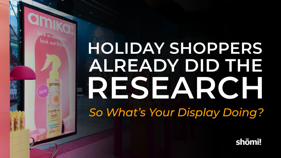 A digital display in a store window features an amika hair product advertisement for in-store holiday shopping, with the text overlay Holiday shoppers already did the research. So what’s your display doing? and the shōmi! logo.